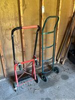 Photo of one green hand truck and one red convertible dolly/cart standing against a wooden wall in shed, showing visible rust and dirt.