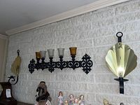 Black metal candle wall sconces with 6 glass candles (3 amber, 3 clear) mounted on white brick wall, also showing a pair of metallic layered fans as wall decor.