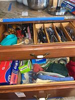 Drawer containing various cooking utensils and kitchen tools including peelers, tongs, knives, forks, and spoons.