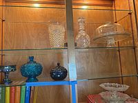 Full view of wood cabinet shelf containing clear cut glass pitcher, decanter, cake stand with cover, carnival glass candy dishes, and frosted serving tray.