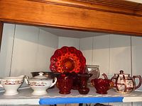 Red glassware and china pieces on a white shelf including round platter, vases, basket bowl, teapot and sugar bowl