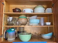 Wooden cabinet shelves with various vintage kitchenware and glass bakeware items including pastel glass bowls, clear glass measuring cups, plastic Tupperware items.