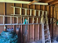 Wooden step ladder and two metal extension ladders hanging on shed wall with other tools and materials below