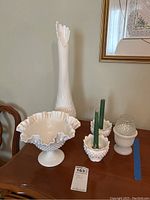 Set of four white milk glass items on wooden surface including tall hobnail vase, candy dish, and two small candle holders, one holding green candles.