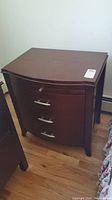Front and side view of the wooden nightstand showing three drawers with metal handles and curved front.