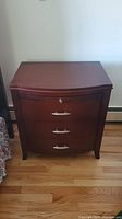Front view showing the three drawer nightstand with curved front, metal handles, and reddish wood finish.