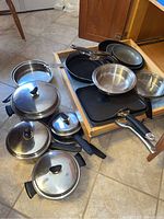 Overview of pots and pans on floor and in open drawer showing variety of metal and non-stick cookware items.
