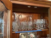 Glassware displayed on a wood shelf including two pitchers, six stemmed glasses, three tumbler glasses, and other glass items