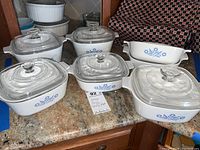 Seven vintage Corning Ware casserole dishes, mostly with clear glass lids, displaying the blue cornflower pattern, arranged on a countertop.