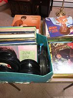 Box and stacks of vinyl records including 12-inch albums and 7-inch singles with visible vinyl sleeves and album covers.