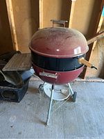 Overall view of the red Weber charcoal grill with round domed lid, black midsection, tripod legs with wheels, and foldable side wooden shelf.