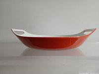 Side profile of the orange enamel paella pan showing the slender cast iron body, white integrated handles with grip cut-outs, and glossy exterior finish.