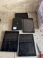 Five tablets arranged on a dirty workbench surface, showing a mixture of brands and models, all powered off and covered with smudges and scratches.