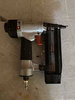 Photo showing a silver and black pneumatic brad nailer with red trigger, lying on a gray floor.