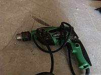 Electric Hitachi hammer drill resting on floor, green and black color with ergonomic grip, power cord coiled loosely.