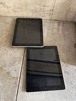 Photo showing two Apple iPads lying on a flat surface, black fronts, silver backs visible, untested condition.