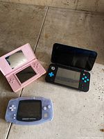 Overhead view showing pink Nintendo DS Lite, black New Nintendo 2DS XL with blue buttons, and translucent purple Game Boy Advance on floor