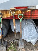 Photo showing a garden pitchfork and square shovel leaning against a rusty red trailer with a tire and white plastic bags visible in the background.