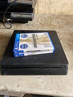 Black PlayStation 4 Slim console with two PS4 games resting on top, Uncharted Collection and FIFA 18, placed on a tabletop with power cable attached.