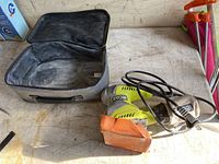 Photo showing the Ryobi electric sander with an orange sanding pad attached, its power cord coiled alongside and a grey soft-sided carry case opened on a work surface.