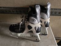 Side and back view of a pair of black and white CCM Tacks ice hockey skates with silver blades, showing lace-up front and reinforced ankle support.