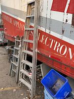 Photo showing multiple ladders leaning against a building with a red and white painted wall. Focus is on a tall aluminum step ladder with 7 steps, showing signs of heavy use and wear.