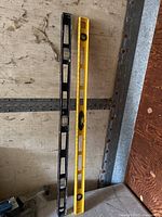 Two levels placed on a worn surface, one yellow and one black, both approximately four feet in length with multiple bubble vials visible for measuring.