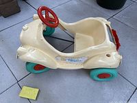 Front angle of ivory/beige plastic toddler pedal car with teal wheels, red steering wheel, and yellow dish sponge for scale.