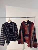 Two vintage ponchos hanging on a rack, showing front side view with the hood and front pouch pocket visible. One is black with multicolor thin stripes, the other features warm tones in patchwork stripe pattern.