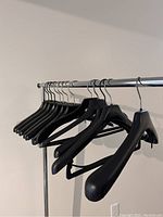 Multiple wide and standard black plastic coat hangers hung on a chrome closet rod. Mainly wide-shouldered hangers at the front, metal hooks attached for hanging. Wall background.