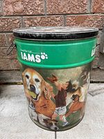 Front and side view of IAMS branded large metal dog food canister showing colorful dog illustrations and green branding band with logo.
