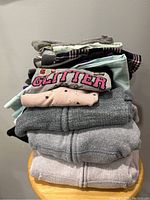 Stack of assorted kids clothing including hoodies, sweatshirts, and shirts with various colors and texts such as 'GLITTER' and star patterns.