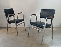 Front angle view of two dark grey vinyl and chrome stacking chairs, showing seat, backrest, armrests with wooden accents, and tubular chrome legs.