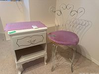 Photo of white bedside table with pink cloth on top alongside white metal chair with pink cushion seat, showing overall lot items