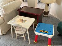 Photo showing the three tables together in a living space with couch and lamp: a Fisher-Price plastic play table with blue top and colorful legs, a white wood child's table with matching chair and colorful bunny illustration, and a dark wood rectangular table with red trim and lower shelf.