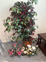 View of large faux leafy plant in wicker pot with small faux flowers on floor nearby.