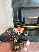 Photo of white donkey figurine, two duck figurines, three raccoon figurines on a base, and metal candle holder with blue candles arranged on black hearth.