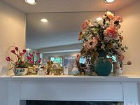 Wide-angle photo showing multiple figurines and planter vases arranged on a white mantel with a mirror background, featuring artificial flowers and decorative ceramic figurines.