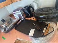 Photo of the entire lot showing electric griddle, coffeemaker box, two electric kettles, and large black speckled baking pan with lid.