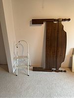 Wooden headboard leaning against wall near small 2-step ladder on carpeted floor
