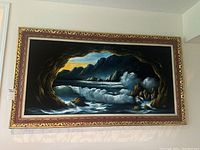 Frontal view of the original velvet painting depicting a seascape inside a cave with crashing ocean waves and distant mountain with sunset sky, housed in ornate gold and red frame.