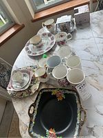 Photo showing multiple Royal Albert Winton floral pattern cups, plates, and mugs arranged on a table, some with visible chips, alongside a black decorative tray with floral design.