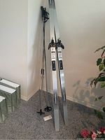 Photo of pair of silver foam fibre cross-country skis with black adjustable bindings standing upright against wall, accompanied by a pair of ski poles with black handles, wrist straps, and round baskets on light carpet.