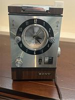 Front view showing Sony vintage radio clock combo with analog clock, tuning, and volume knobs, plus AM/FM switch. Wood accents and brushed metal exterior visible.