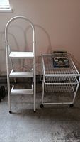 Photo showing the metal folding step stool next to the rolling metal cart with wire basket drawers, both items white metal, placed on a concrete floor