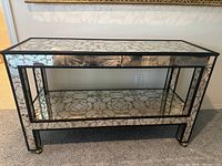 Full view of rectangular glass design table with black metal frame and wheels, mirrored glass surfaces showing cracks and antique silver pattern.