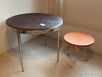 Two round tables in a room, one large with dark brownish metal top and metal legs, one small orange with ornate metal legs.