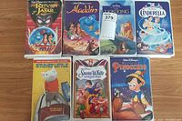 Front covers of seven Disney VHS tapes including Return of Jafar, Aladdin, The Lion King, Cinderella, Stuart Little, Snow White, and Pinocchio laid flat on a wood surface.