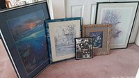 Five framed pieces of art leaning against a white door on carpet. Includes different styles and colors, large to small sizes.