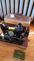 Overview of black Singer sewing machine set on wooden base with power cord and blue thread spool. Tan hard carrying case behind machine. Green box labeled Singer in front, presumably containing accessories.
